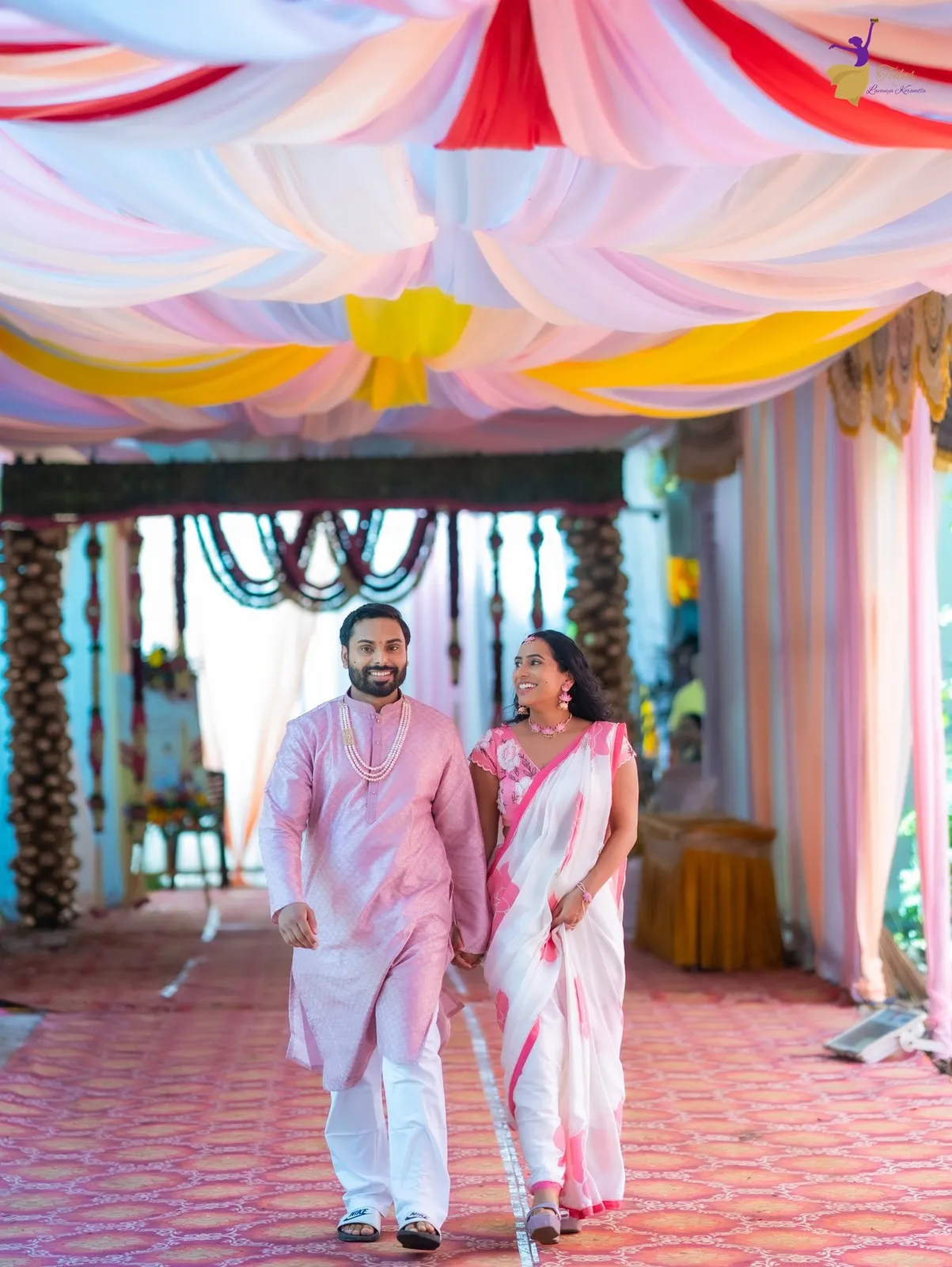 Manoj and Priya in pastel pink and white walking under a colourful haldi canopy, Telugu wedding Amalapuram – TBLK