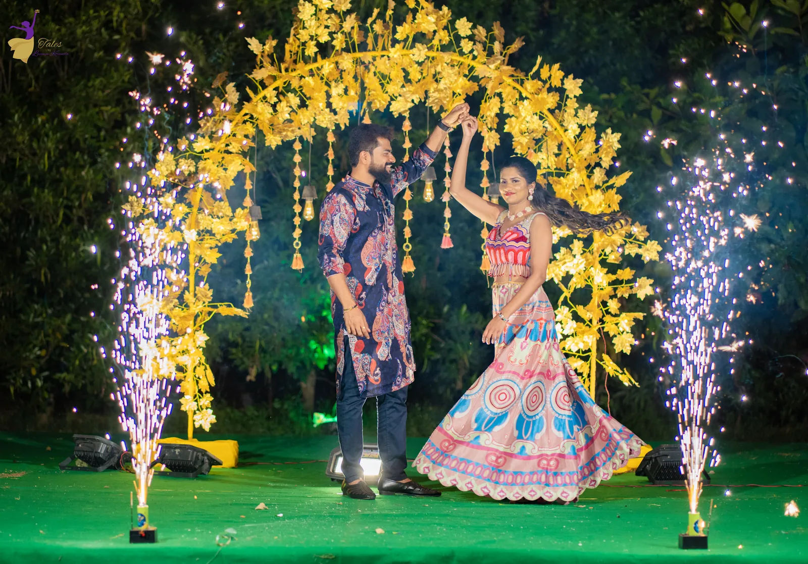 Wide shot of Krishna and Vishwasa in festive prints under a golden floral arch with cold sparklers on both sides