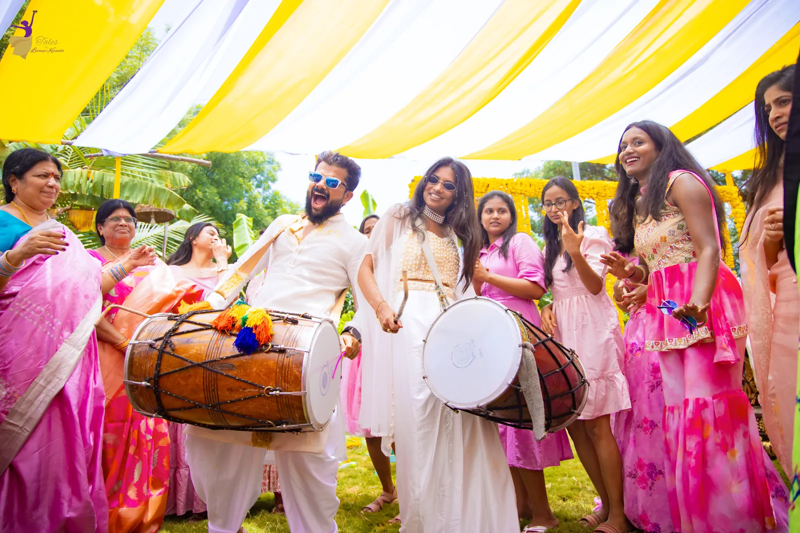 Wide shot of the couple and cousins dancing as two dhol players drum under yellow and white haldi drapes