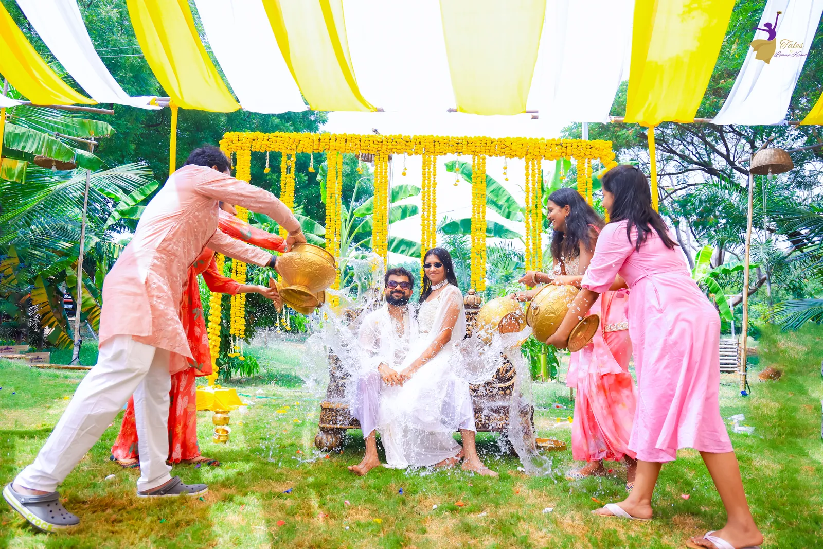 Wide shot of family pouring water from brass pots over the seated couple under yellow and white haldi drapes
