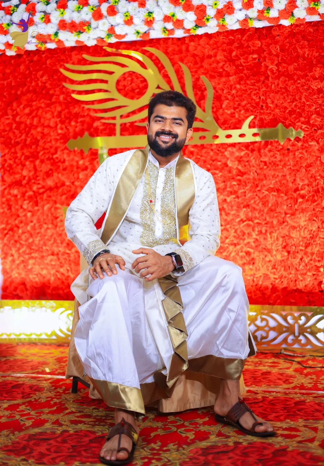 Krishna in white and gold seated before a wall of red roses with a golden peacock-feather motif at his ceremony