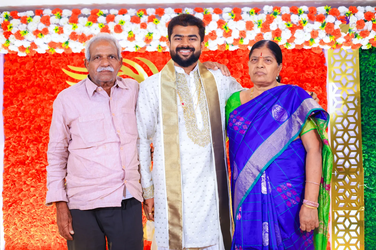 Krishna in white and gold with family before the red rose wall at his ceremony