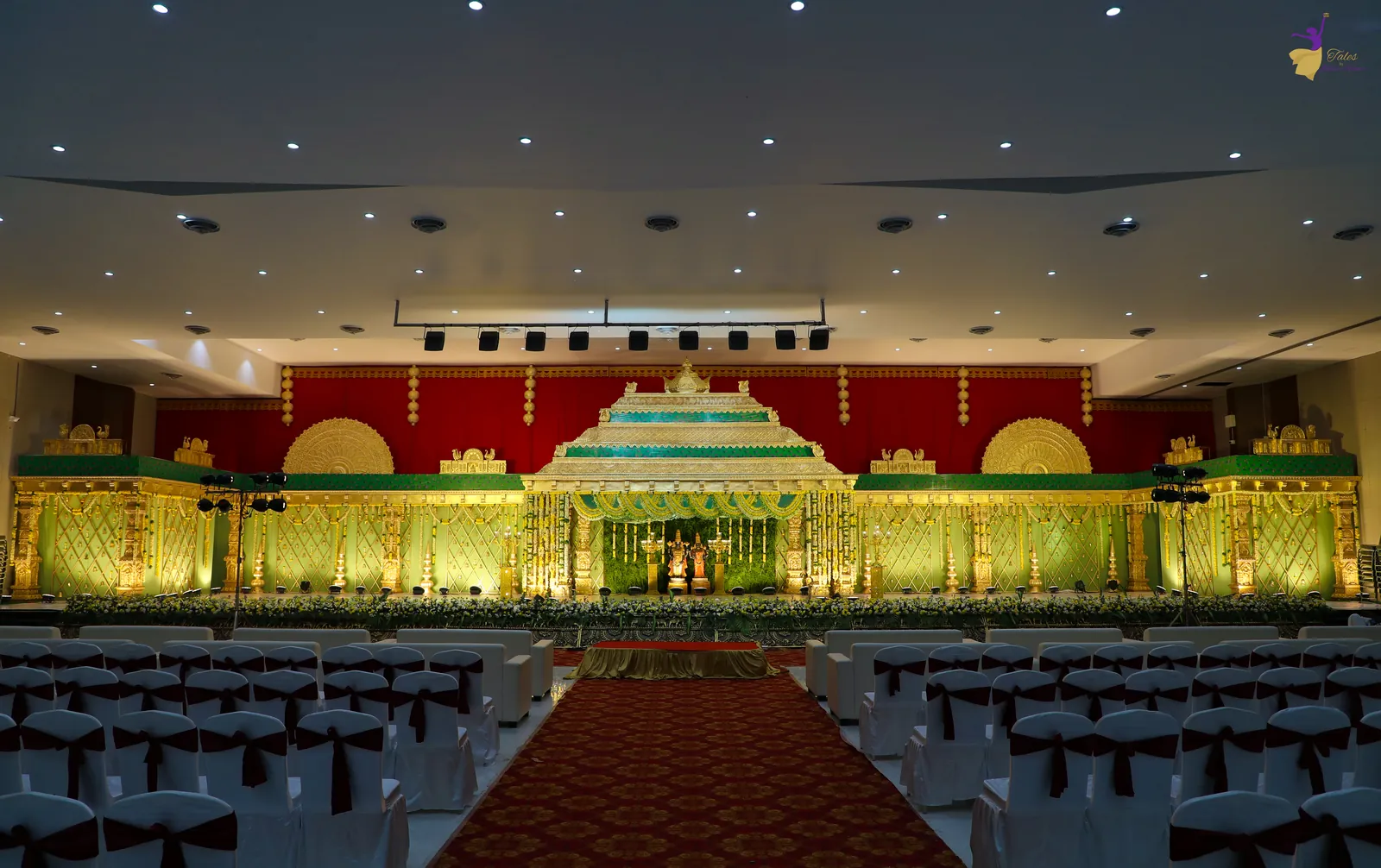 Wide view of the grand wedding hall with a golden mandap stage, red carpet aisle and rows of seating