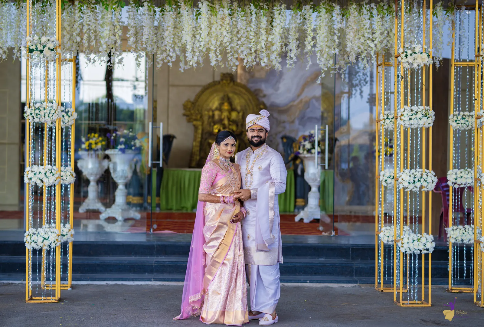 Wide shot of the bride and groom under a white floral arch at the venue entrance