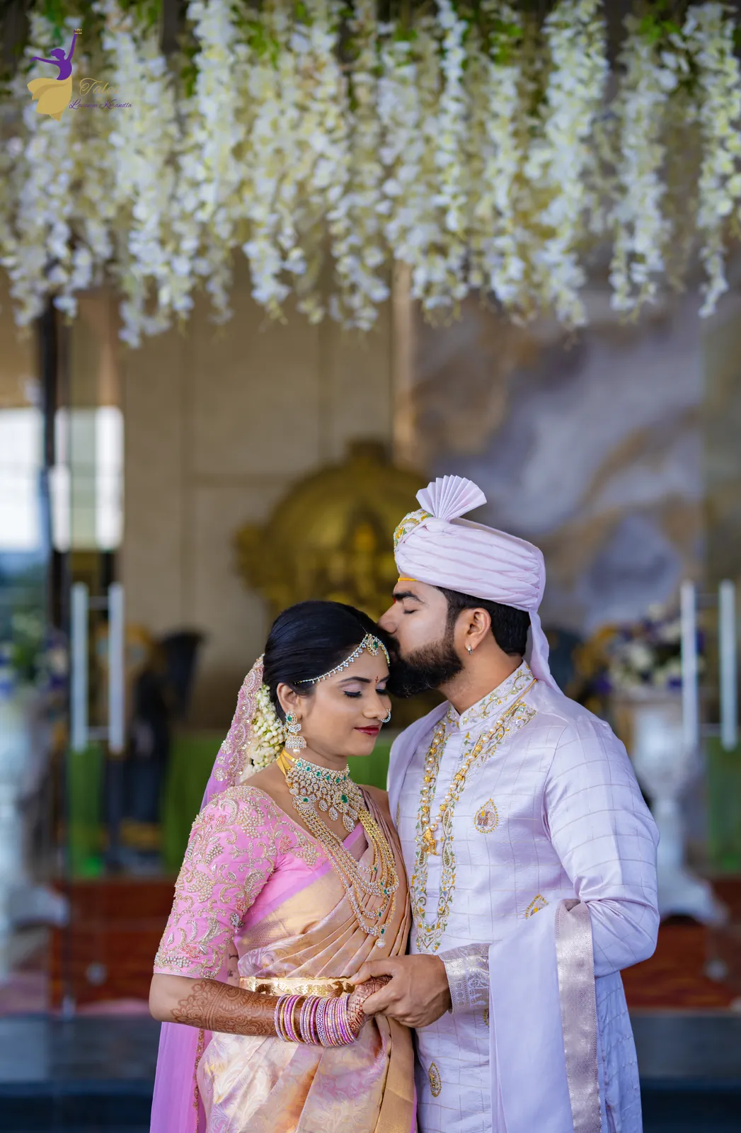 The groom kissing the bride’s forehead under hanging white flowers at the venue