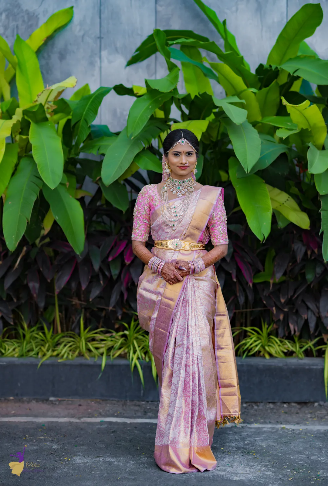Full-length bridal portrait in a pink and gold silk saree before tropical leaves