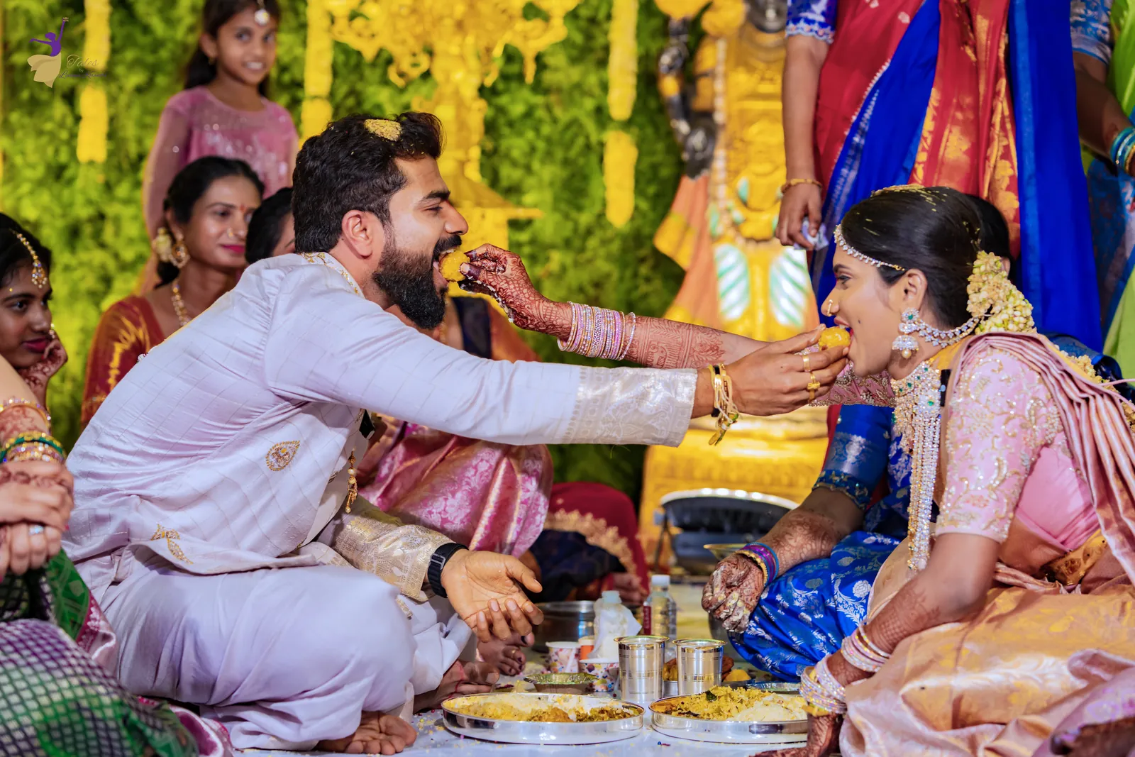 The groom feeding the bride during a ritual on the mandap, with family and guests all around