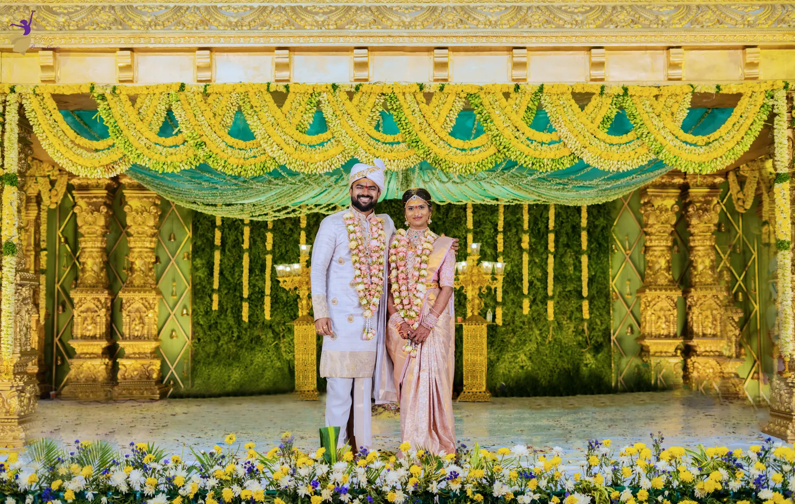 Wide view of the ornate gold and green mandap with the bride and groom standing at its centre