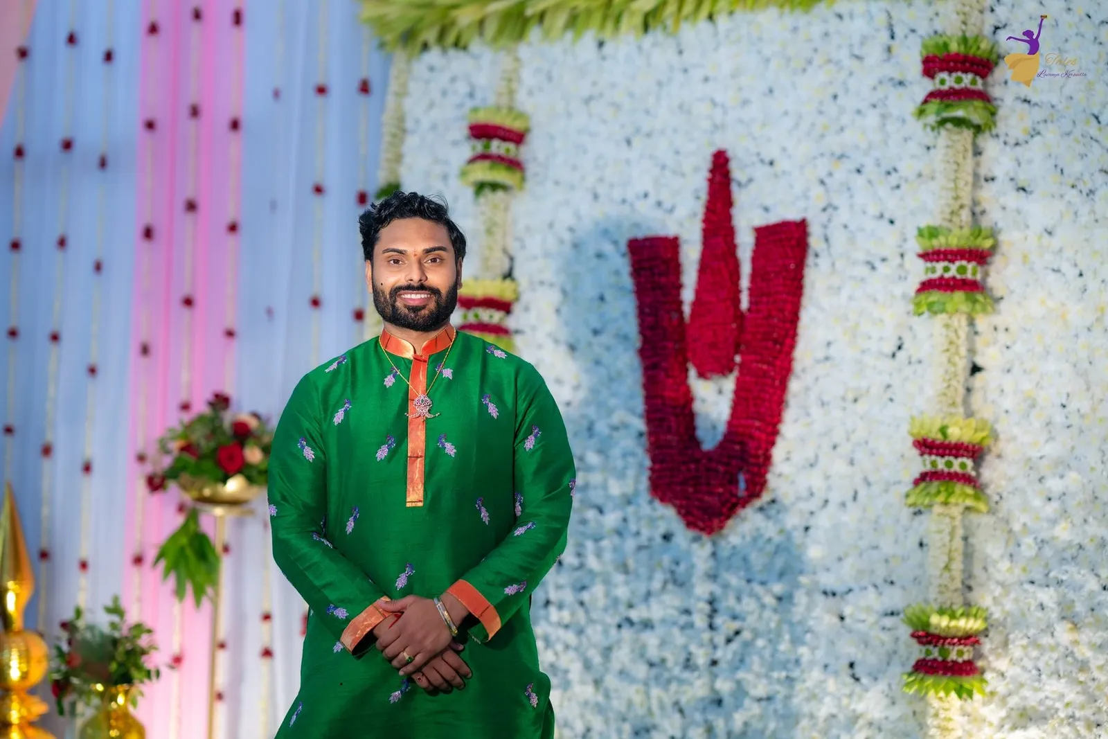Manoj in a green kurta smiling before a white floral wall with a red namam motif at his Pellikoduku – TBLK