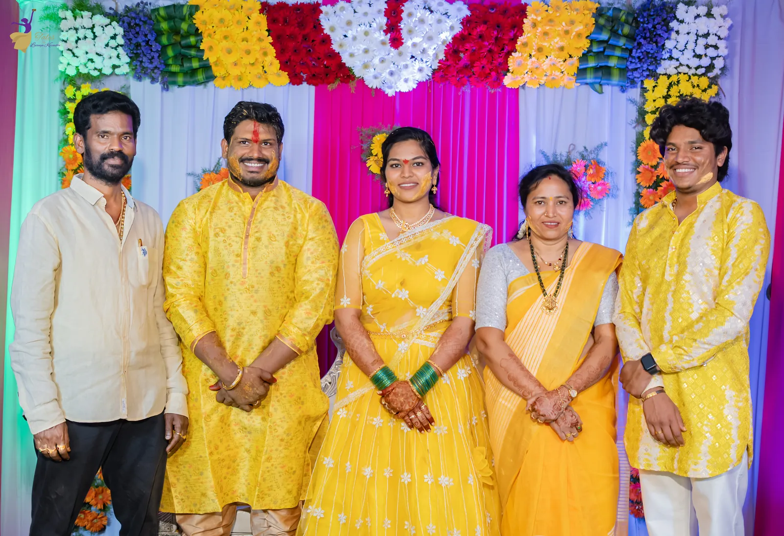 Ravinder and Anuhya in yellow with their cousins in front of a colourful flower wall at the haldi
