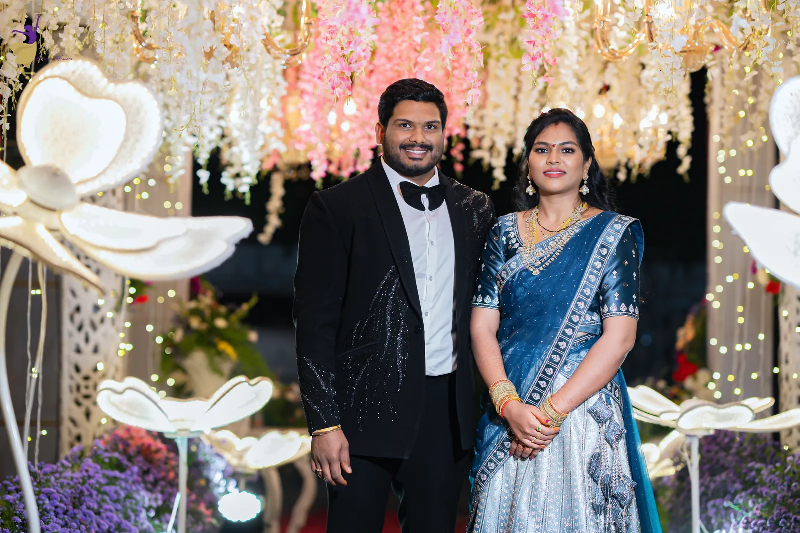 Couple portrait of Ravinder in a black tuxedo and Anuhya in blue among white flower sculptures