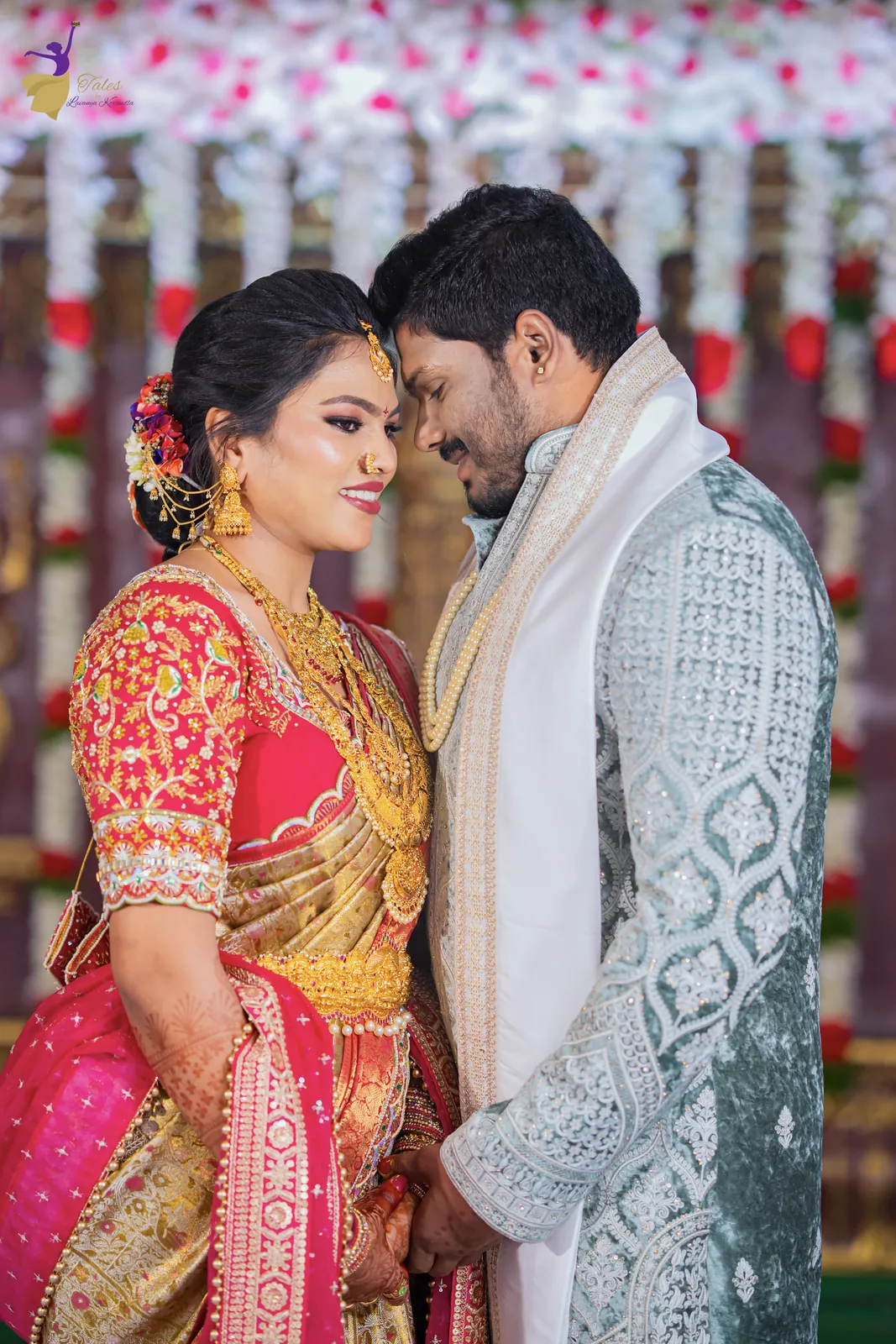Ravinder and Anuhya leaning in close in wedding silks before a red and white flower curtain