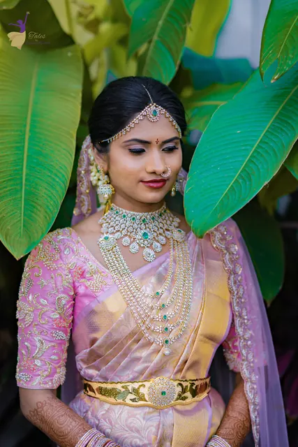 Bride in pink and gold silk saree with layered emerald and diamond jewellery among banana leaves