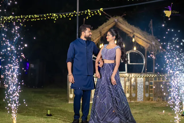 The couple in navy and lavender standing before a lit thatched gazebo with cold sparklers on both sides