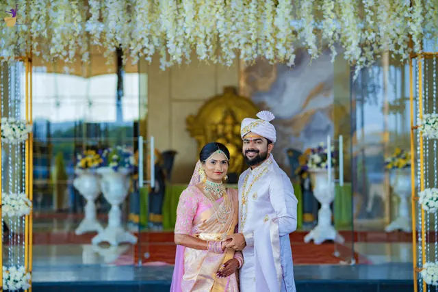 Wide shot of the bride and groom under a white floral arch at the grand venue entrance