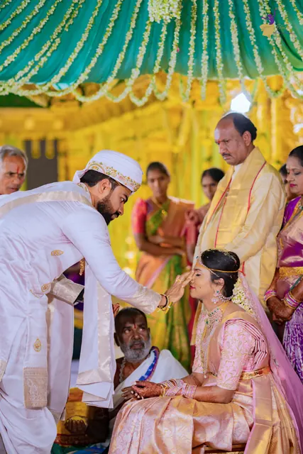 Wide shot of the groom tying the thali around the bride’s neck as family look on under the mandap