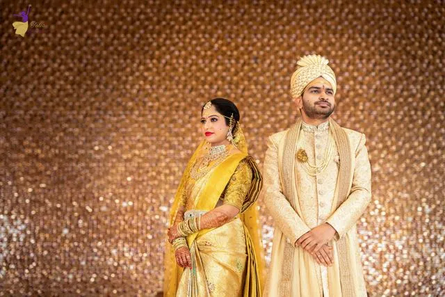 Wedding couple in gold and cream attire against a shimmering bronze backdrop, Hyderabad