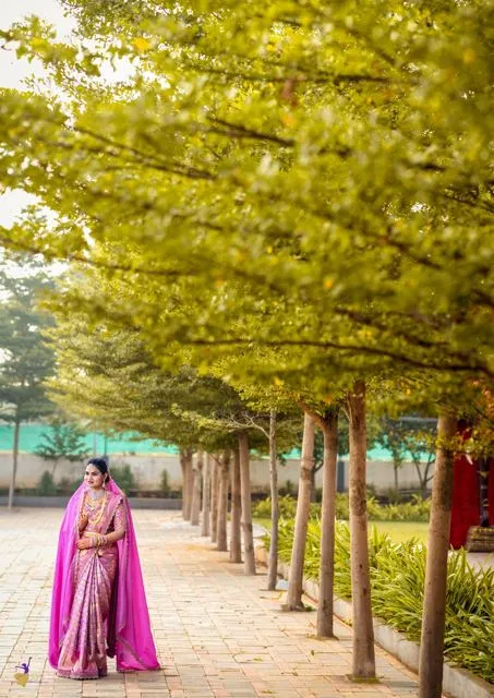 Bride in pink and purple saree walking a tree-lined path